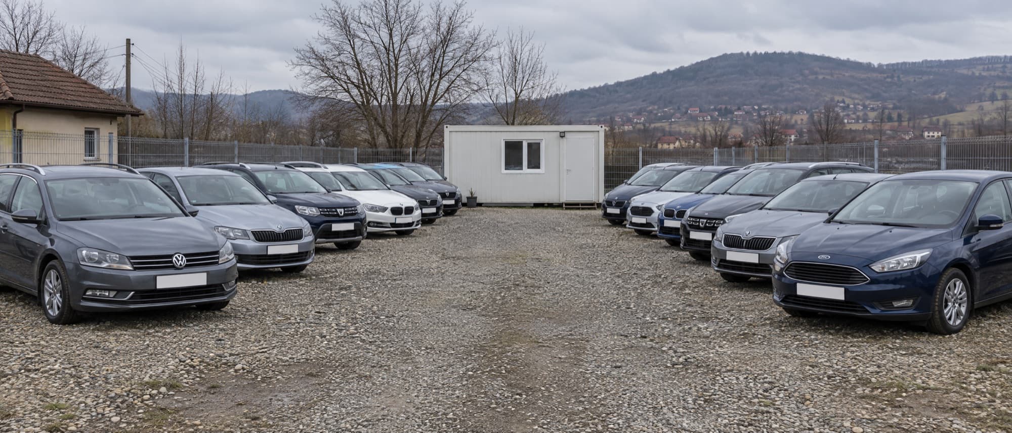 Un parc auto second-hand: două rânduri de mașini pe pietriș și un birou mic, la marginea unui oraș.